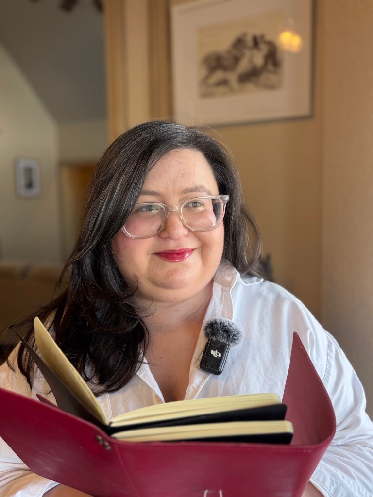 An image of a white woman with long brown hair and glasses holding a journal and smiling